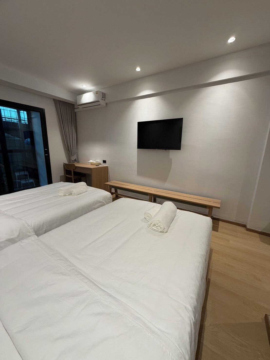 A hotel room with two beds, a desk, a flat-screen TV, and an air conditioning unit.