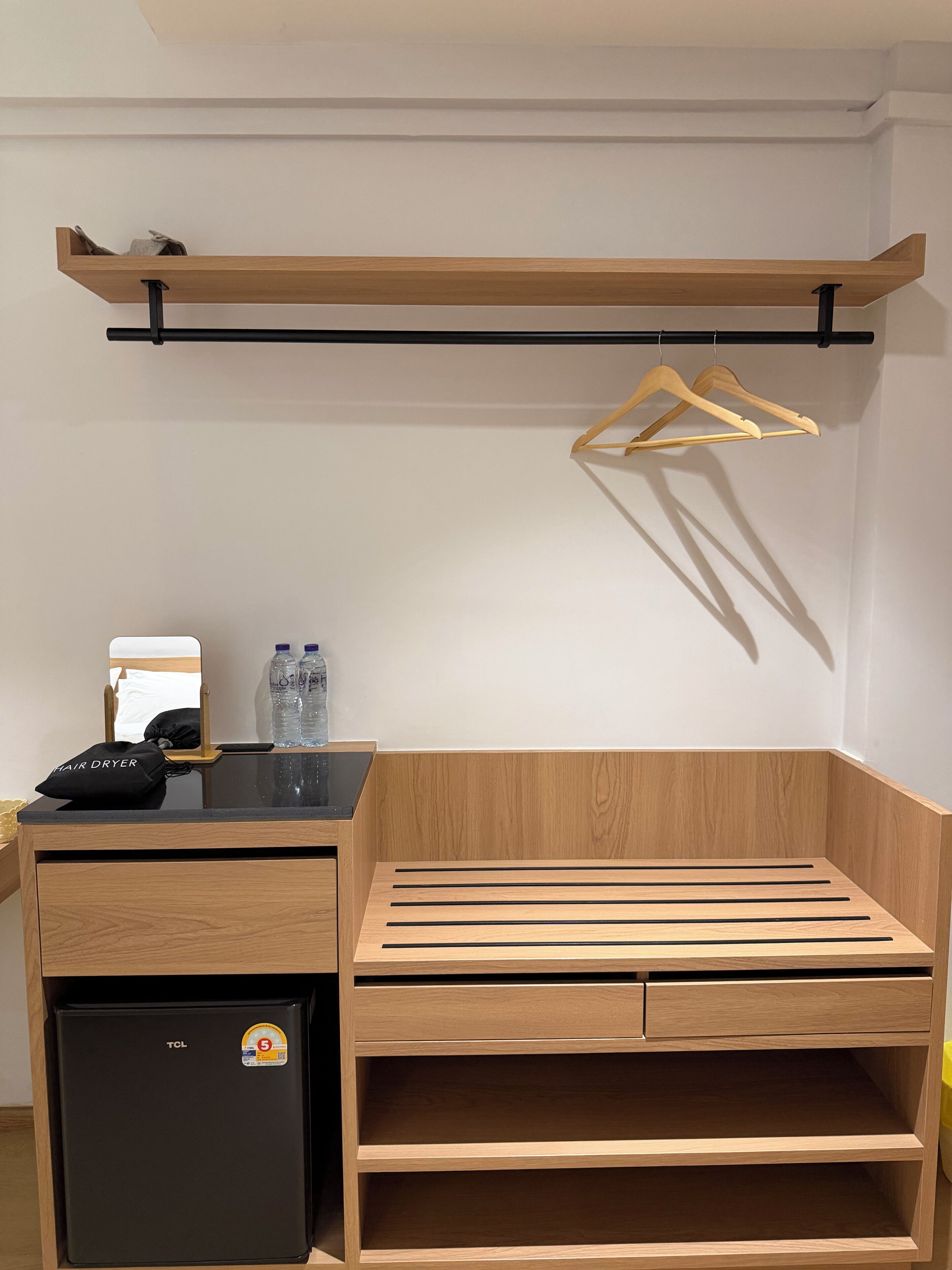 Ein kleiner Raum mit einem Holztisch, einem schwarzen Kühlschrank, einem Spiegel und einer Kleiderstange.