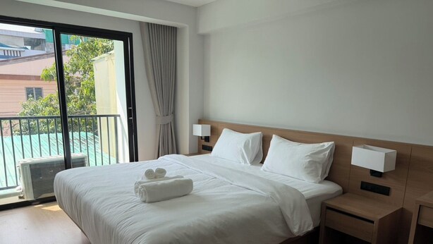 A hotel room with a large bed, a bedside table, a nightstand, a window with a view of buildings, and a balcony. - 611 Hotel (Phetchaburi)