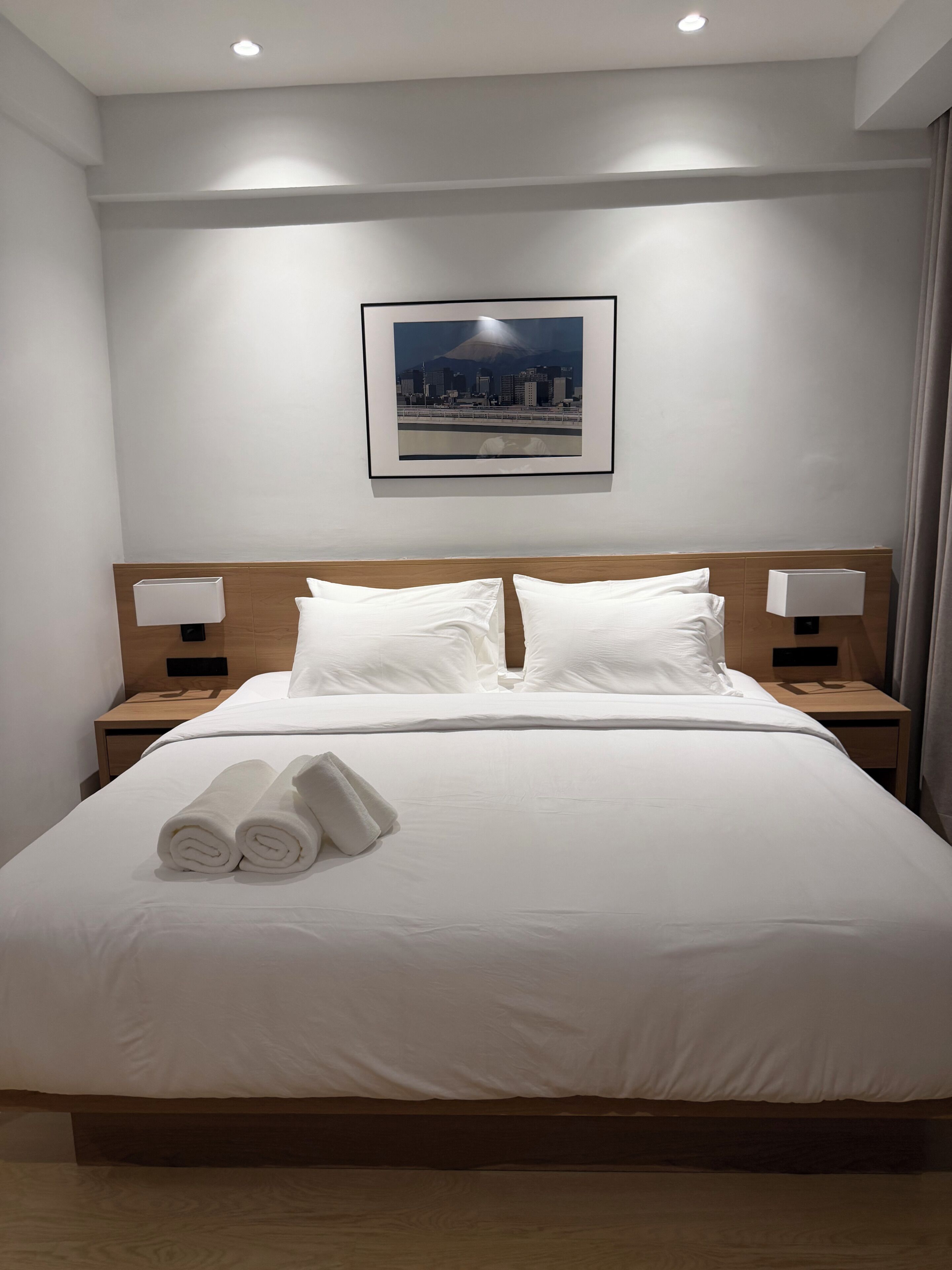 A hotel room with a neatly made bed, two bedside tables with lamps, a framed picture on the wall, and a pair of rolled towels on the bed.