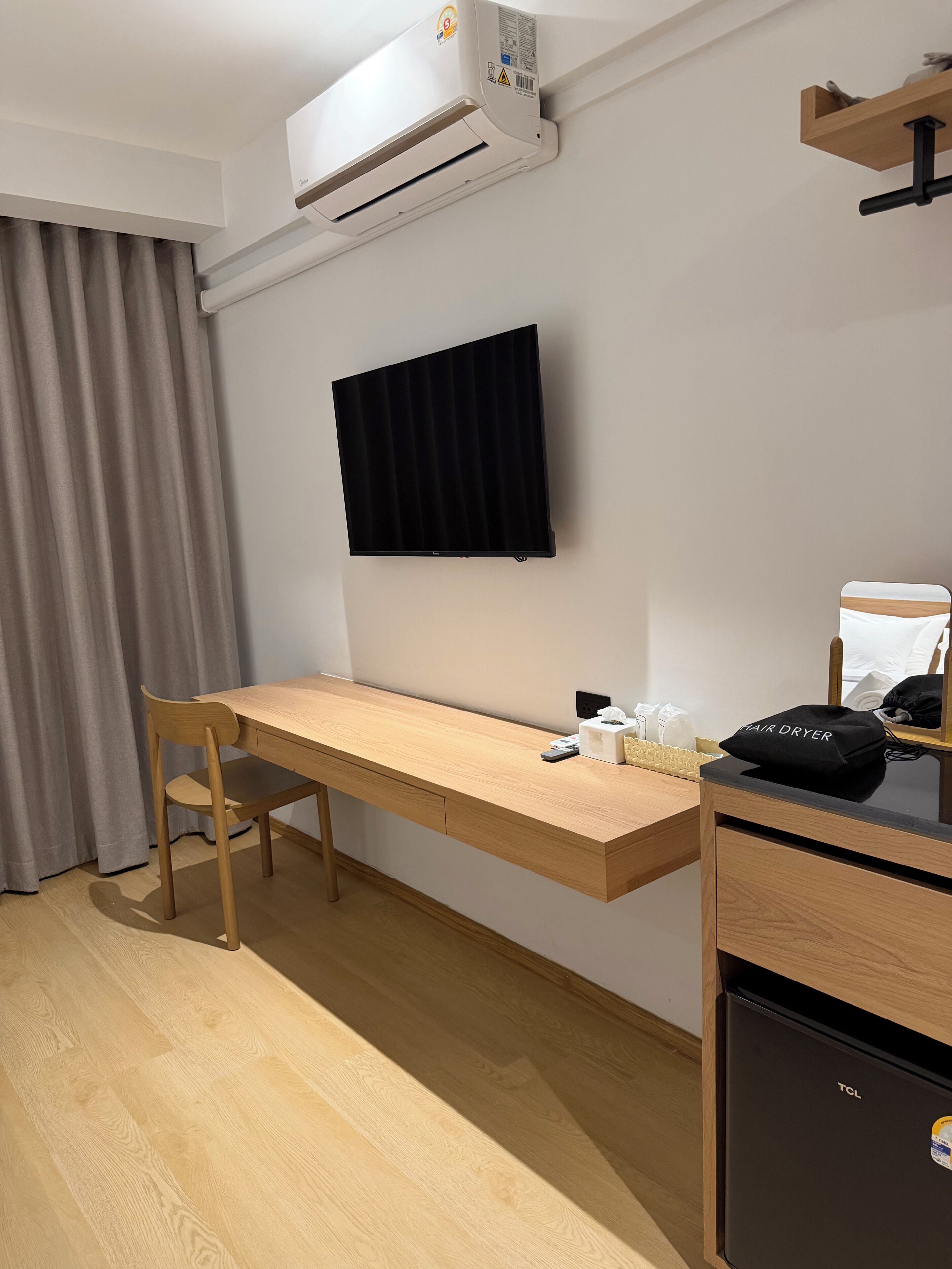 A room with a wooden desk, a chair, a TV mounted on the wall, a hair dryer on a small table, and a mini fridge.