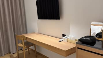 A room with a wooden desk, a chair, a TV mounted on the wall, a hair dryer on a small table, and a mini fridge.