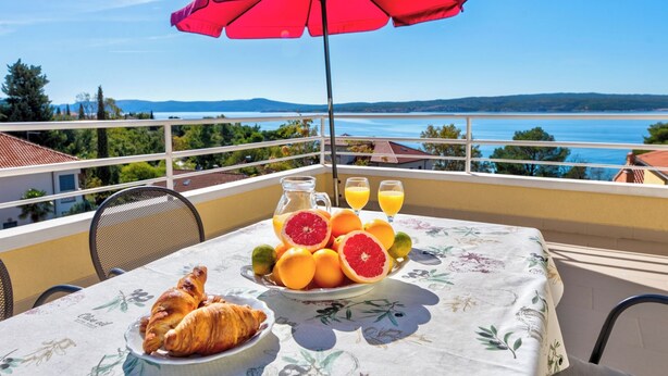 En balkong med ett bord dukat för frukost, ett rött parasoll och en vacker utsikt över havet.