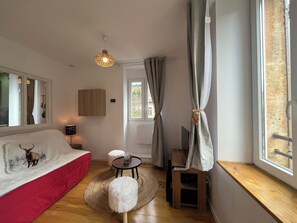 A modern living room with a red sofa, a round wooden table, a small stool, a ceiling light fixture, and a window with curtains. - Cosy, refurbished apartment in gated residence. (La Bourboule)