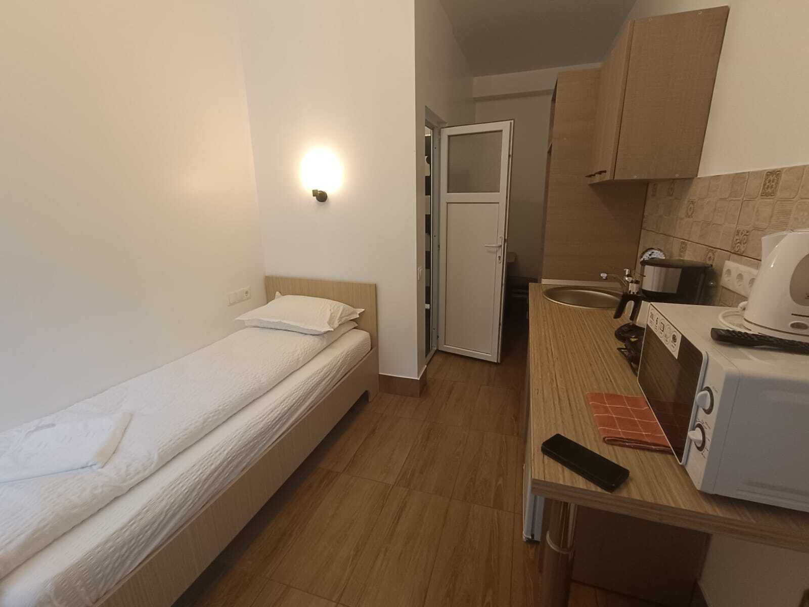 A small, compact room with a single bed, a kitchenette with a microwave and toaster, and a door leading to a bathroom.
