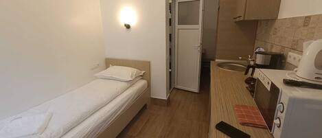 A small, compact room with a single bed, a kitchenette with a microwave and toaster, and a door leading to a bathroom.
