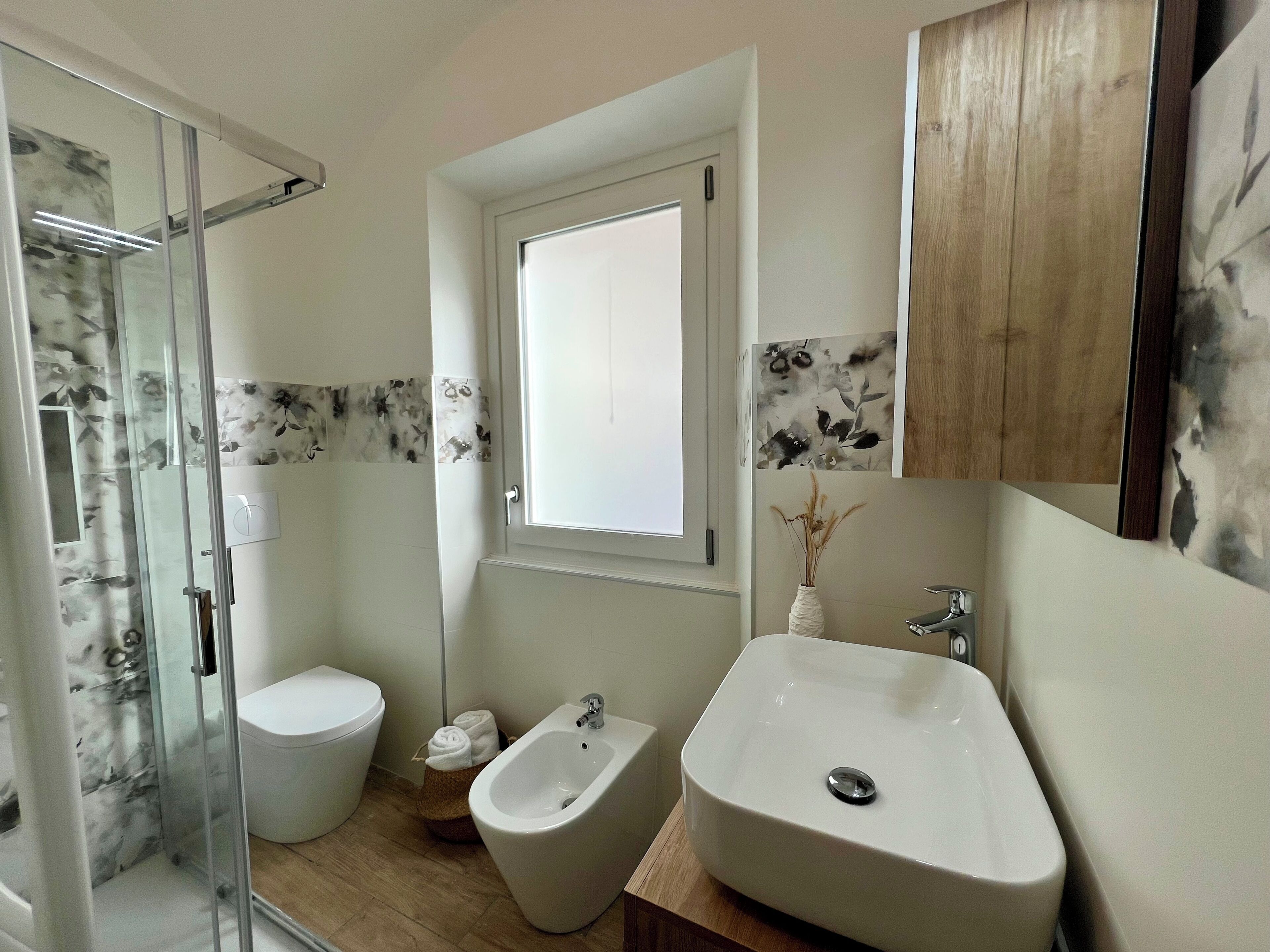 A bathroom with a glass shower enclosure, a toilet, a bidet, a sink, and a window.