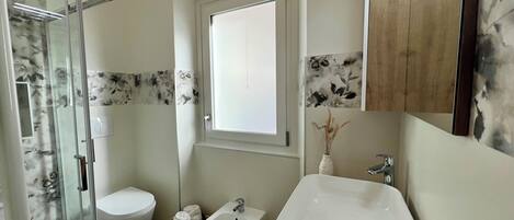 A bathroom with a glass shower enclosure, a toilet, a bidet, a sink, and a window.