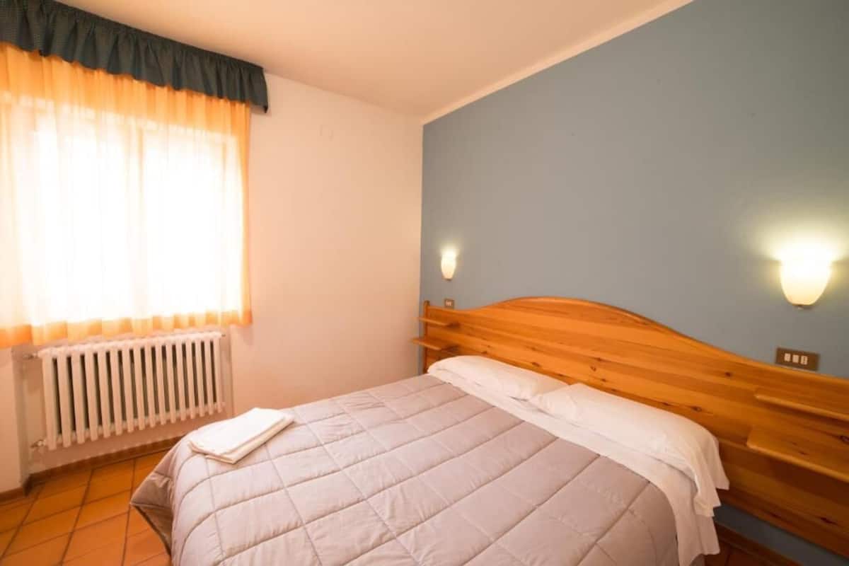 A bedroom with a wooden headboard, a bed with white bedding, a window with sheer curtains, a wall-mounted light, and a radiator.