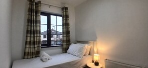 Ein Einzelbett mit weißen Laken, eine Nachttischlampe und Vorhänge mit Karomuster an einem Fenster.