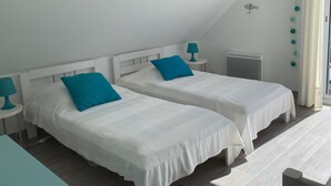 Zwei Einzelbetten mit weißer Bettwäsche und blauen Kissen, ein kleiner Tisch mit einer Lampe, eine Heizung und ein Fenster mit Vorhängen.