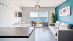 Un saló modern amb un sofà, una taula de menjador, una televisió i una vista al mar a través d'una porta de vidre.