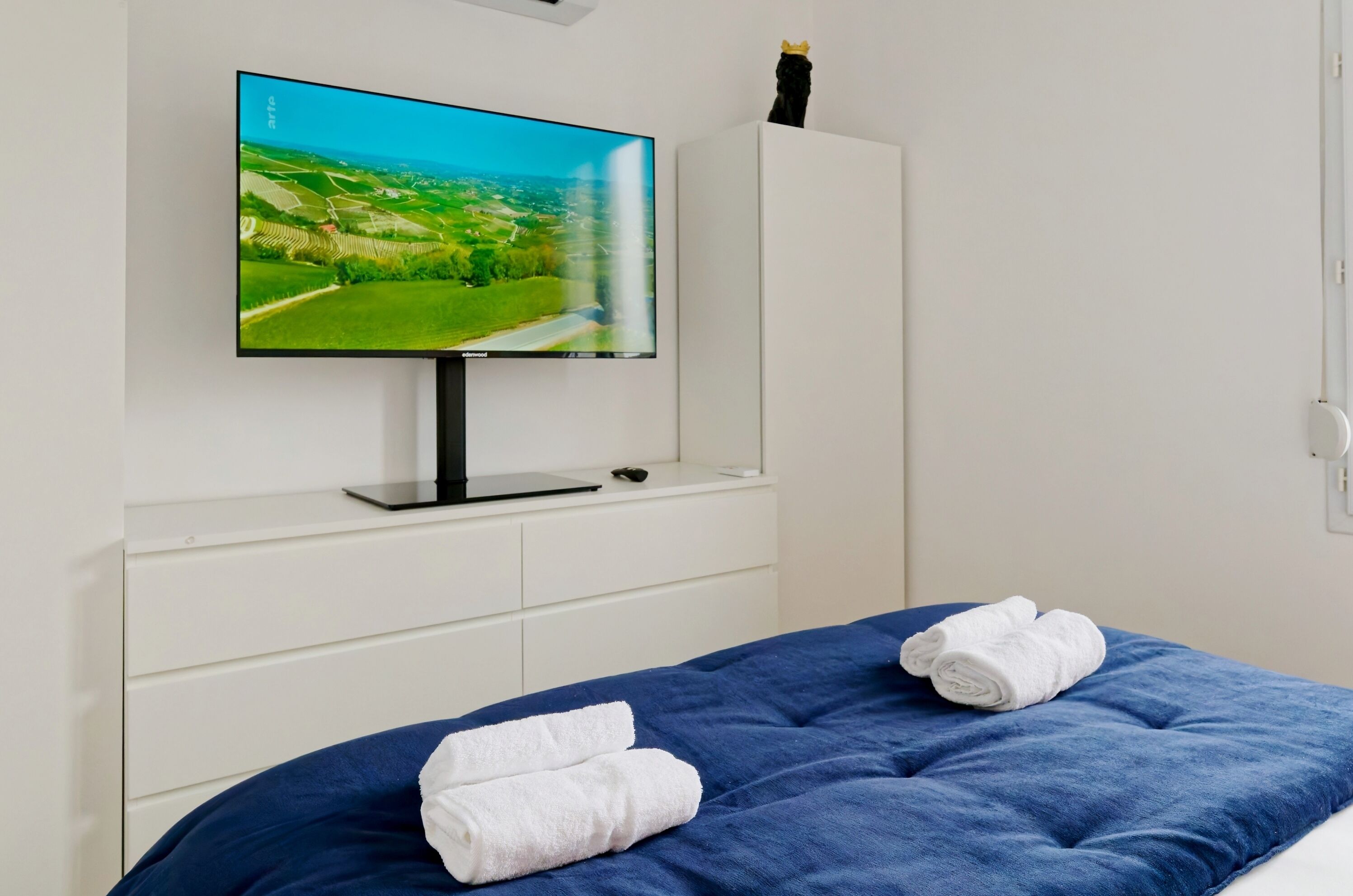 A bedroom with a TV on a white cabinet, a bed with blue bedding, and two rolled towels.