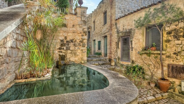 A stone courtyard with a small pool, potted plants, and traditional stone buildings.