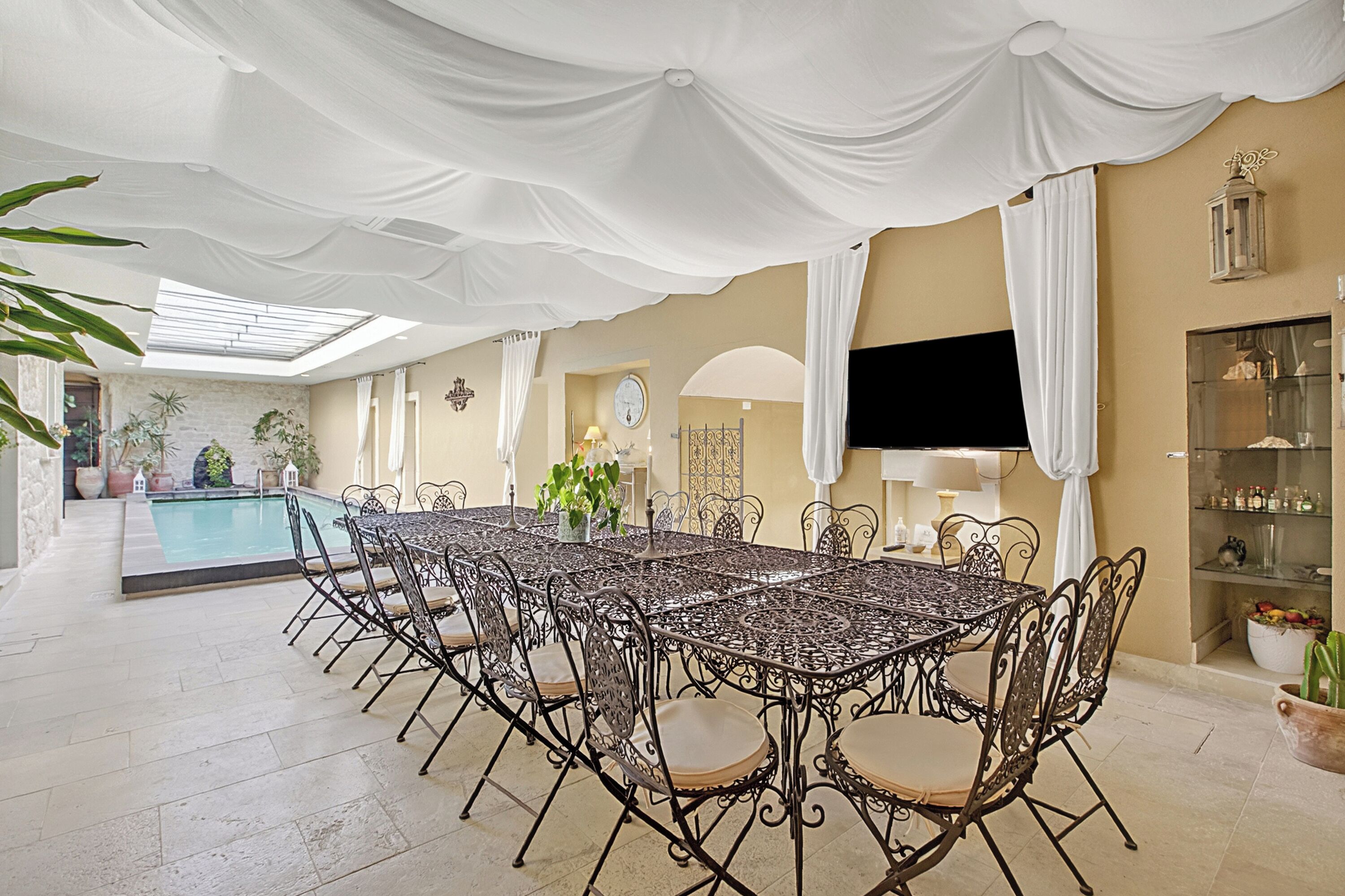 Un espace repas avec une piscine, des rideaux blancs et un téléviseur.