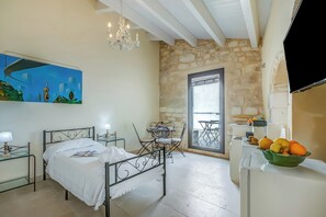 A bedroom with a bed, bedside tables, a painting, a chandelier, a stone wall, a balcony with a table and chairs, and a refrigerator with fruit.
