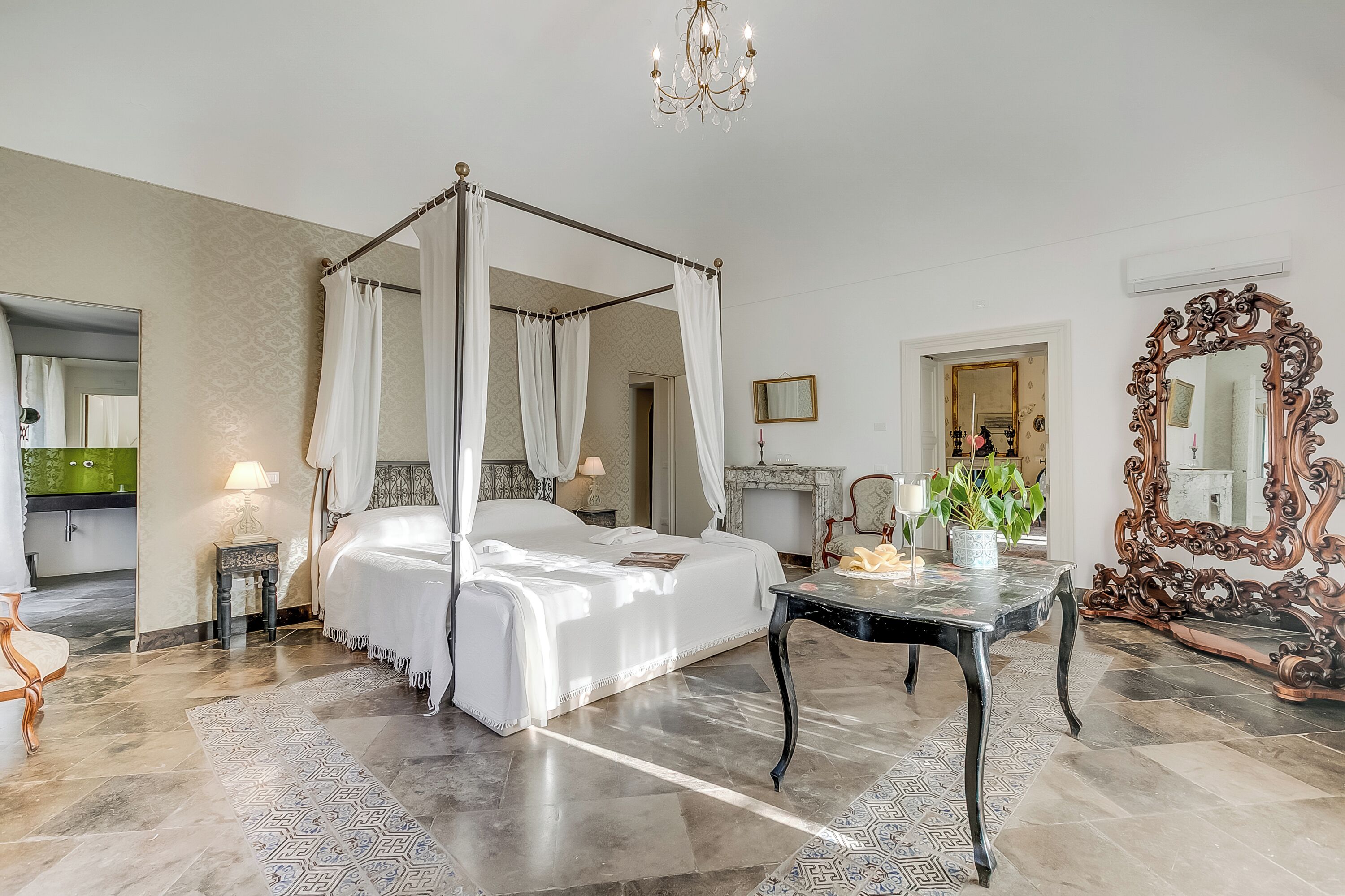 A four-poster bed, a chandelier, a mirror, and a dining table in a hotel room.