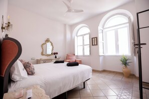 Een slaapkamer met een bed, een stoel, een kaptafel, een spiegel, een kroonluchter en een groot raam.