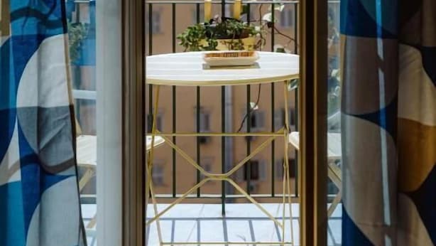 Un balcon avec une petite table et des plantes en pot, donnant sur un immeuble doté de nombreuses fenêtres.