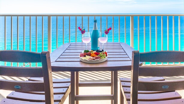 Et træbord med et frugtfad og to glas med en blå drik, placeret mod en balkon med udsigt over havet.
