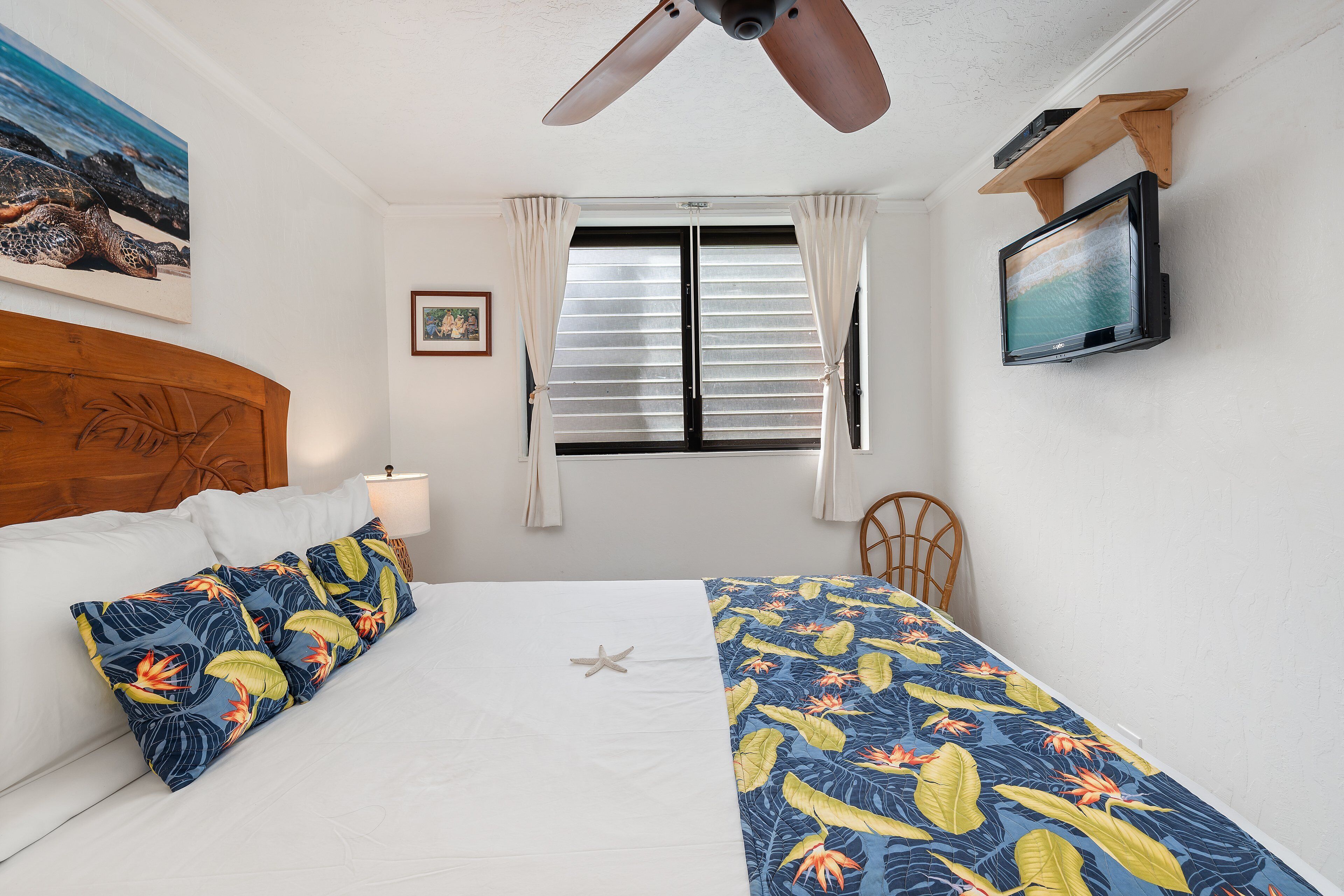 Een slaapkamer met een bed, een plafondventilator, een televisie, een raam met gordijnen en een schilderij aan de muur.