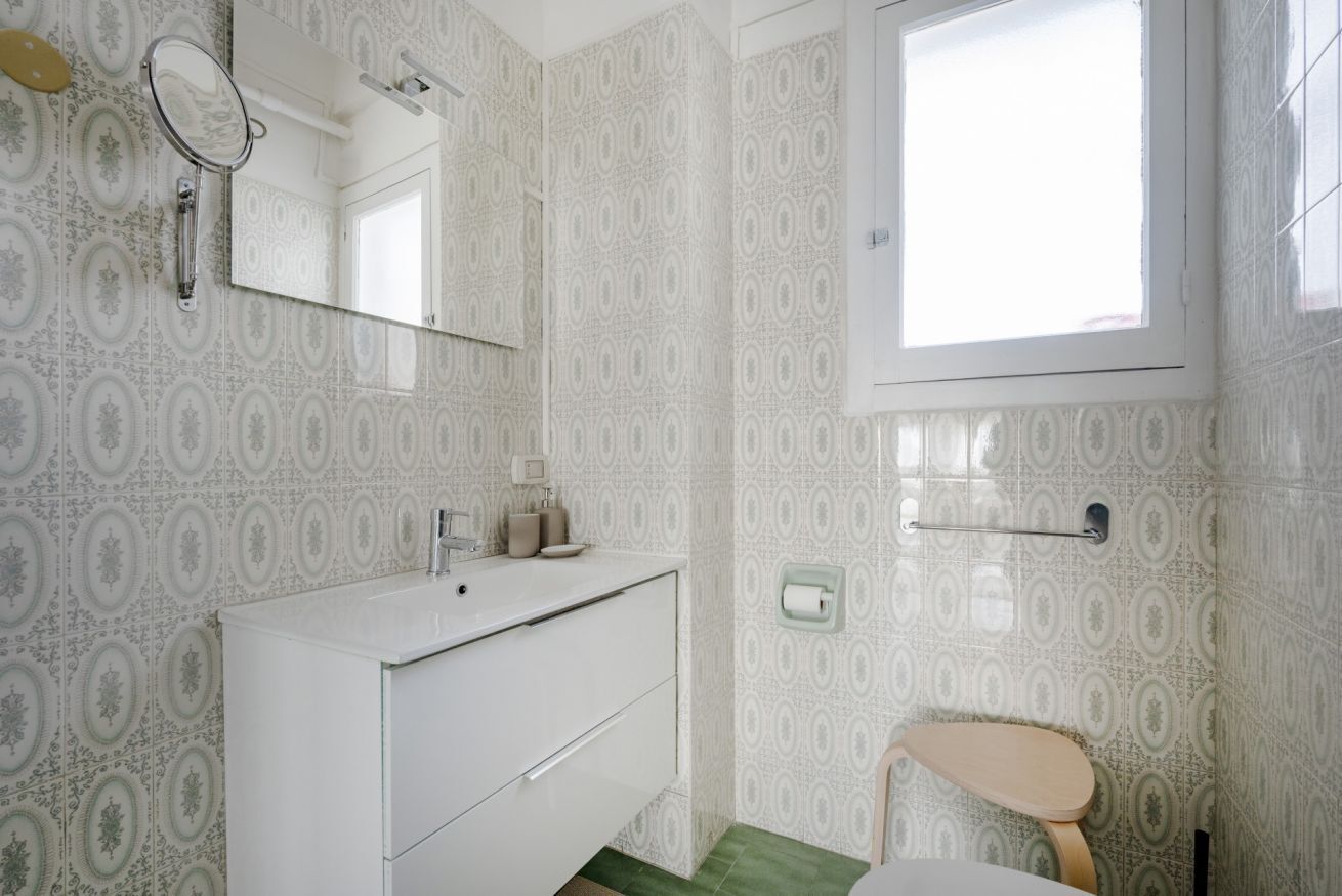 A bathroom with patterned wallpaper, a white sink, a mirror, a toilet, and a window.