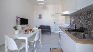 Et moderne køkken med hvide skabe, et spiseområde med et hvidt bord og stole, en grå sofa og et fjernsyn på et hvidt skab.