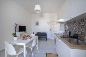 A modern kitchen with white cabinetry, a dining area with a white table and chairs, a gray sofa, and a TV on a white stand. - GuestHost - Dimora Buonvino (Bari)