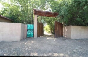 Une entrée fermée par un portail, surmontée d’une arche en bois, qui mène à un sentier bordé d’arbres.