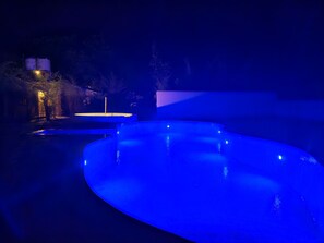 A nighttime swimming pool with blue lighting, surrounded by trees and a building with illuminated water tanks.
