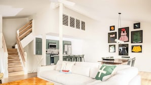 A modern living room with a wooden staircase, a white sofa, a wooden coffee table, and a dining area with chairs. - The Family Clubhouse at Abbey Springs (Fontana)