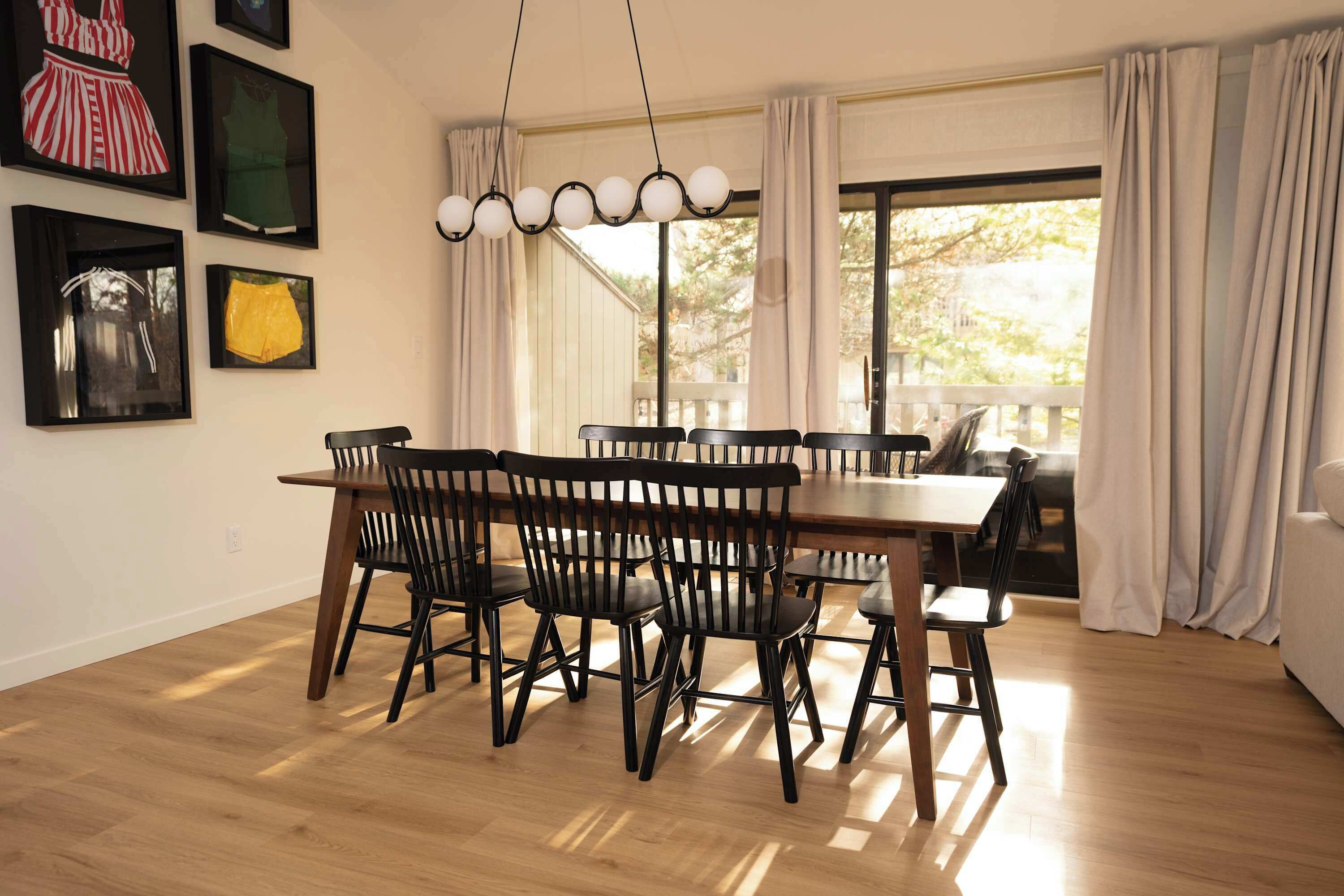 A dining area with a wooden table and chairs, framed artwork on the wall, and a view through a large window.
