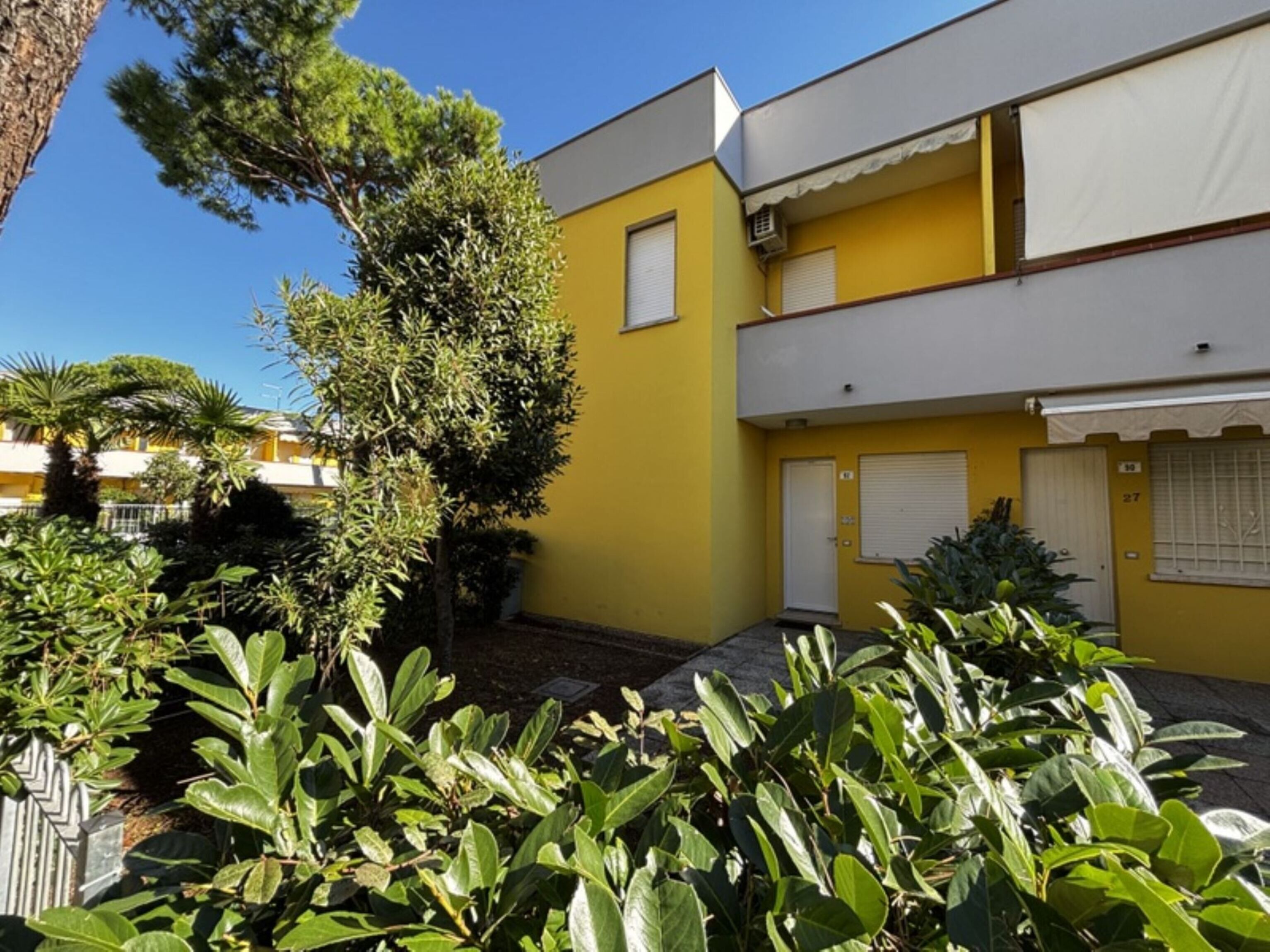 A yellow building with a white door, surrounded by greenery and a clear blue sky.