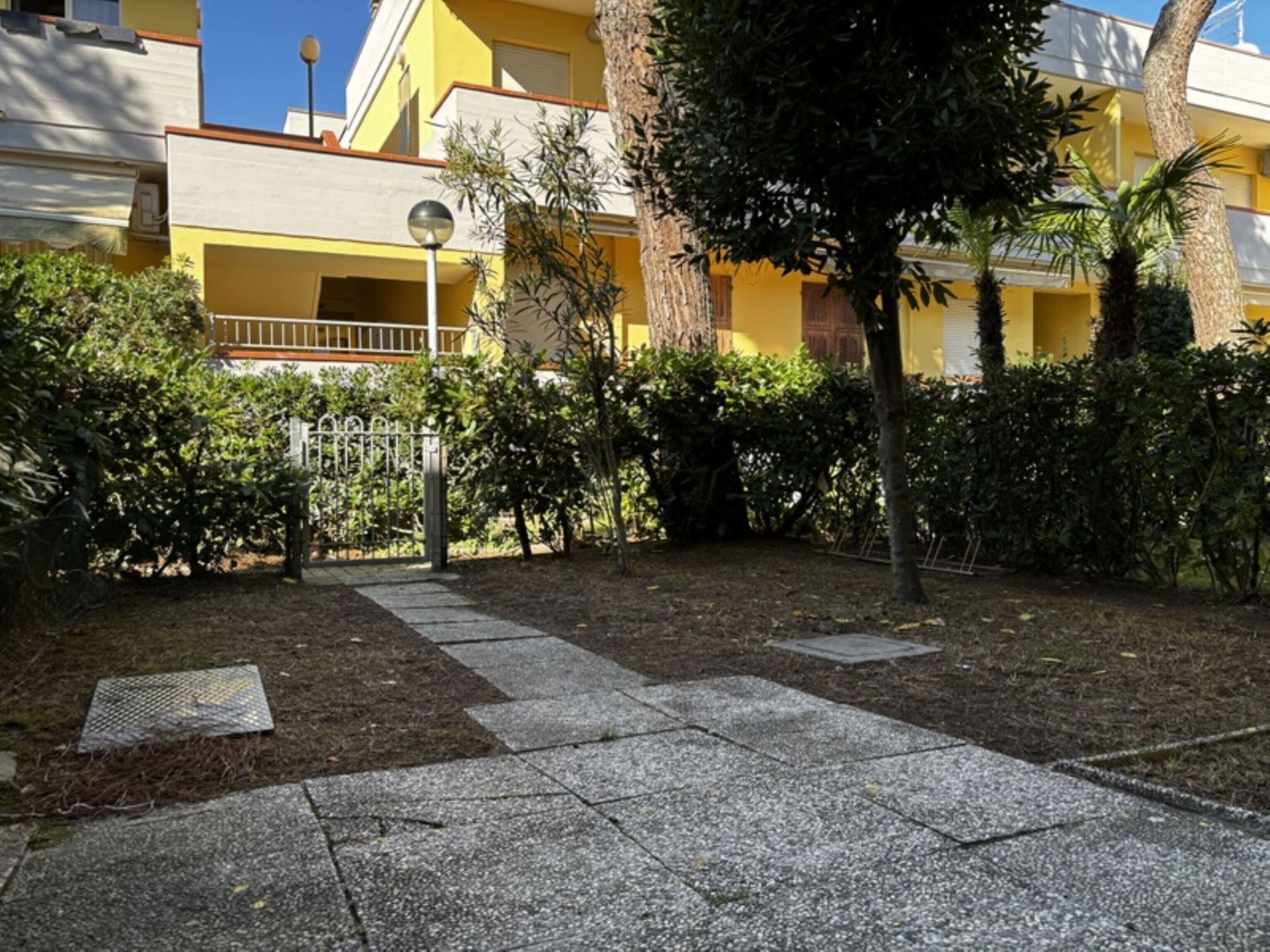 A courtyard with a paved walkway, a metal gate, and a yellow building with balconies.
