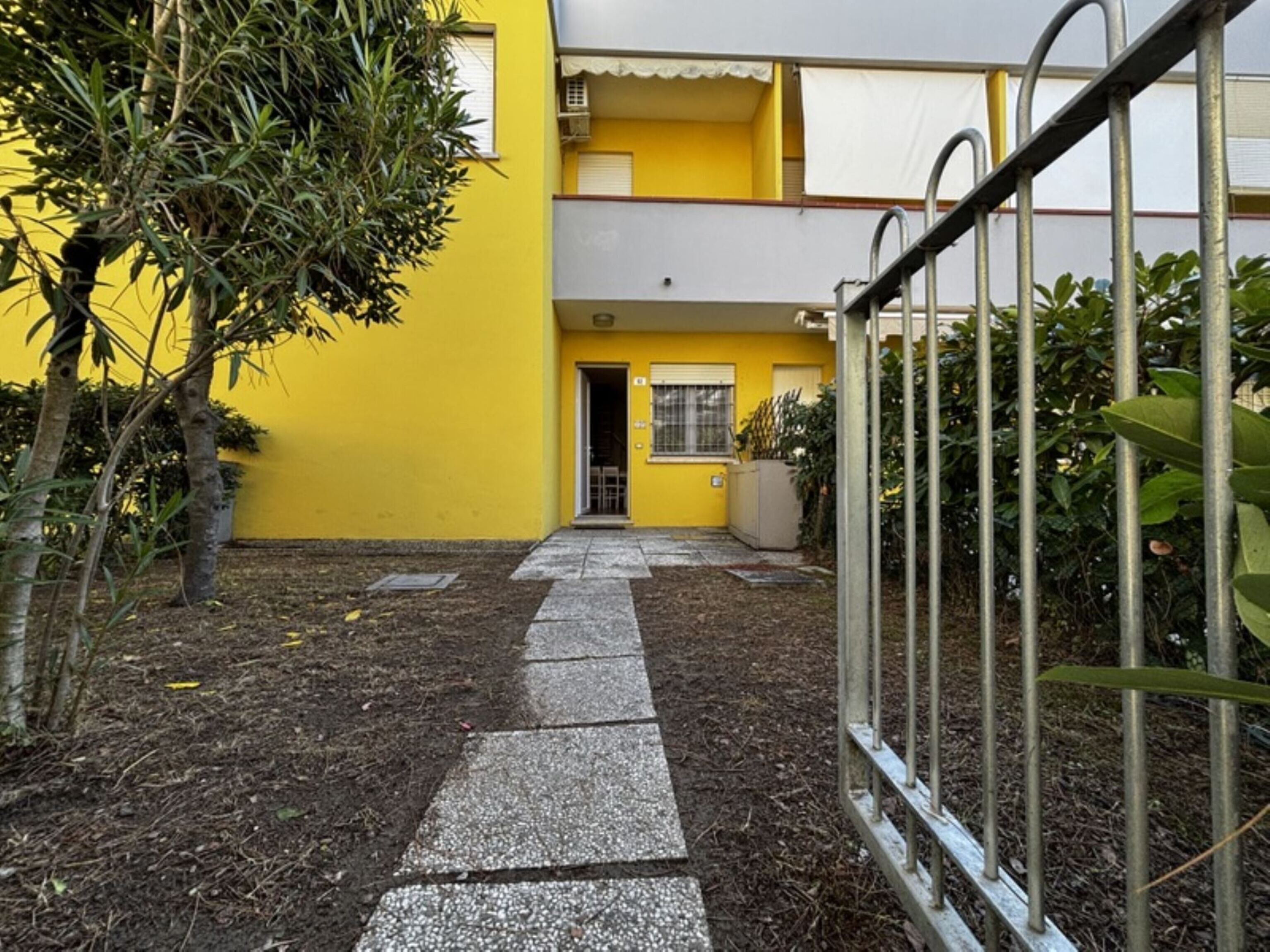 A yellow building with a paved walkway leading to a door, surrounded by greenery and metal fencing.
