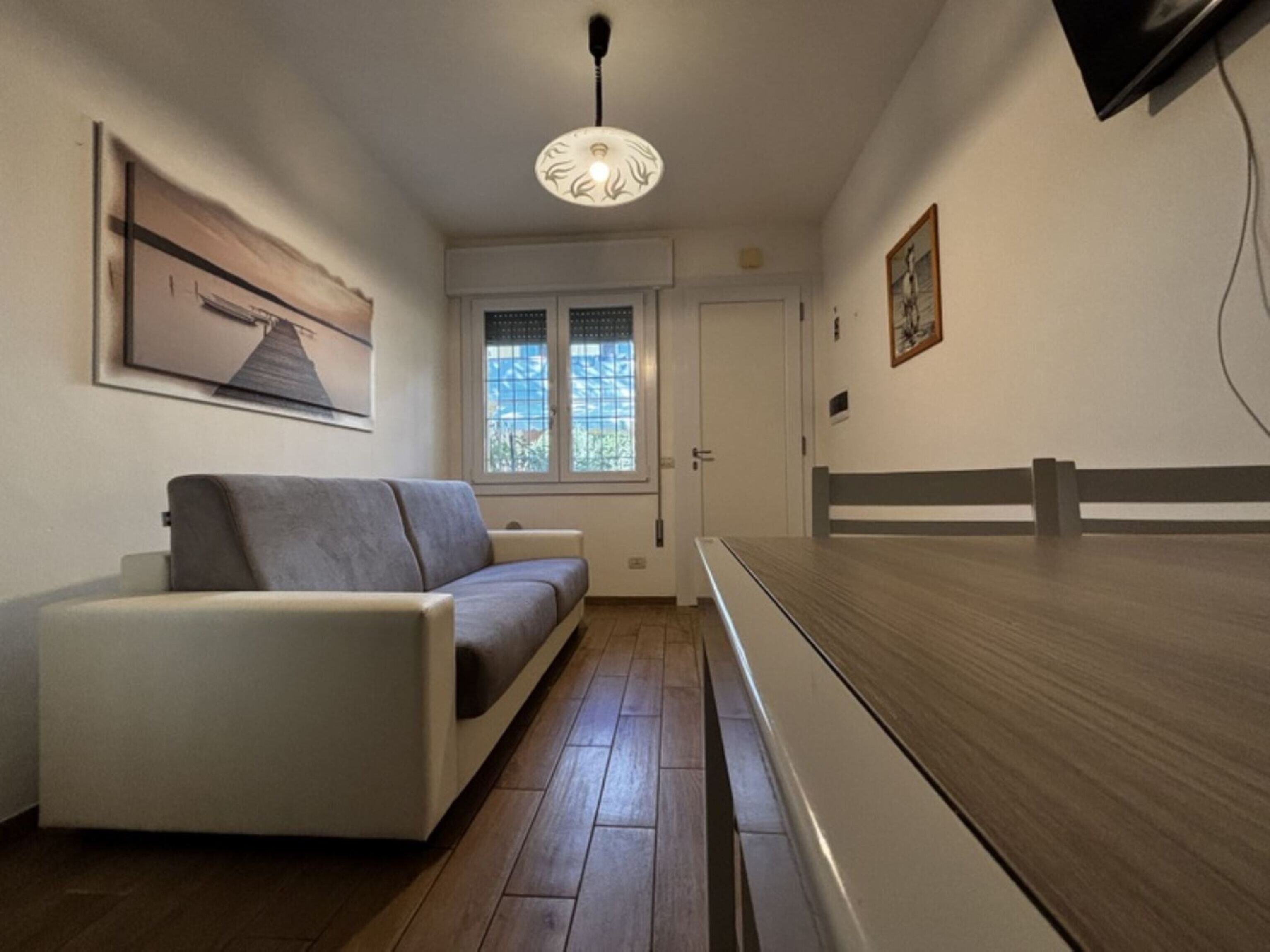 Quarto moderno com sofá, piso de madeira, luminária no teto, janela com persianas e porta.