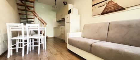 A living room with a sofa, a dining table with chairs, a kitchen area, and a staircase.
