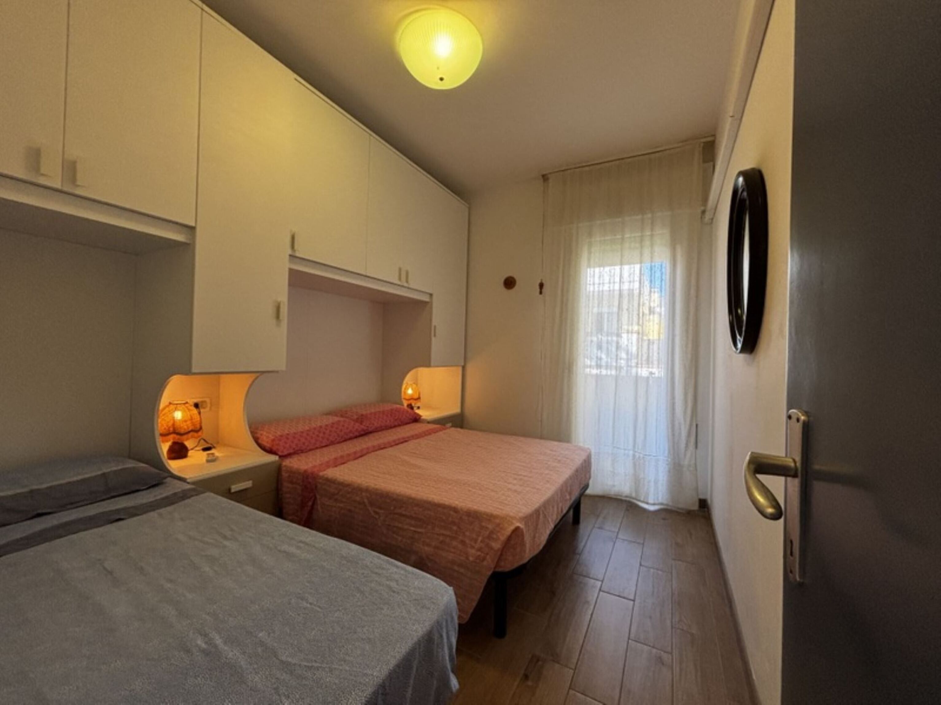 Quarto pequeno e bem iluminado com duas camas, um guarda-roupa, mesa de cabeceira com luminárias e uma porta com puxador.