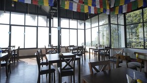 A spacious indoor dining area with large windows, wooden tables and chairs, and colorful prayer flags hanging from the ceiling. - Longitude Homestay (Kalimpong)