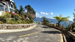 A road with a stone wall on one side and a building with a balcony on the other. - Longitude Homestay (Kalimpong)