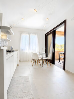 Eine moderne Küche mit weißen Schränken, ein Essbereich mit einem Glastisch und weißen Stühlen sowie eine Glasschiebetür, die zu einer Außenpatio führt.