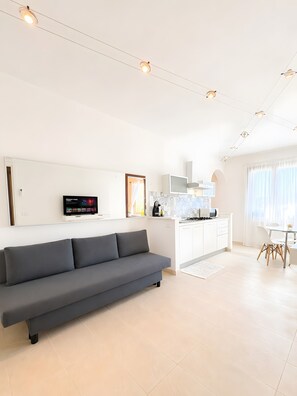 Ein modernes Wohnzimmer mit einem grauen Sofa, einem Flachbildfernseher und einer Küchenzeile mit weißen Schränken sowie einem Esstisch.