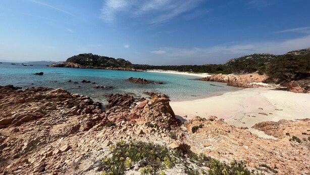 Un paisaje costero con terreno rocoso, aguas turquesas cristalinas y una playa de arena.