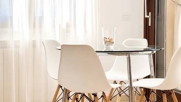 Un espace de salle à manger moderne avec une table en verre, des chaises blanches et une vue sur le paysage urbain à travers des rideaux transparents.