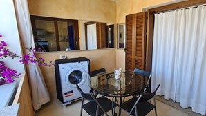 Un balcón pequeño con lavadora, una mesa de vidrio y sillas, y vista a la cocina a través de un espejo.