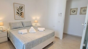Una cama bien tendida con almohadas, dos lámparas de noche, una obra de arte con hojas enmarco y una puerta a la derecha.