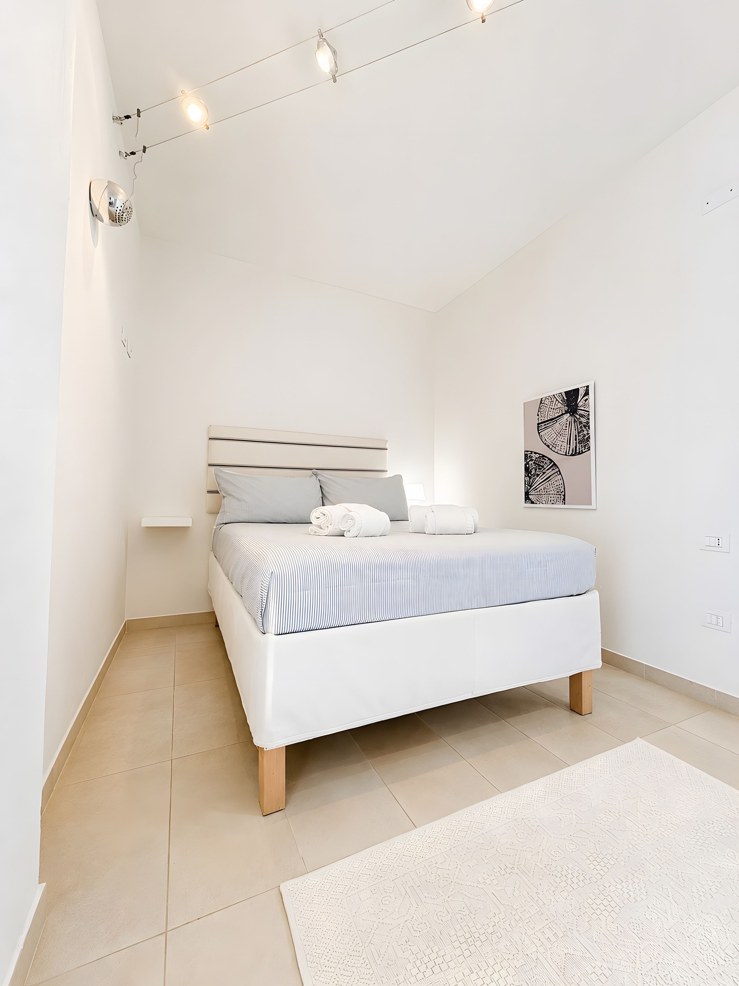 A minimalist bedroom with a bed, a bedside table, a wall-mounted light fixture, and a framed artwork.