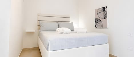 A minimalist bedroom with a bed, a bedside table, a wall-mounted light fixture, and a framed artwork.