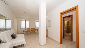 A spacious living area with a sofa, dining table, and chairs, a large pillar, and a wooden door leading to a bathroom. - Apartment 'A 104 Puntavilla' – Perfect for Large Families (La Maddalena)