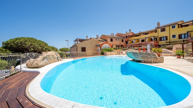 A swimming pool with a wooden deck, surrounded by buildings with Mediterranean architecture. - Apartment 'A 104 Puntavilla' – Perfect for Large Families (La Maddalena)